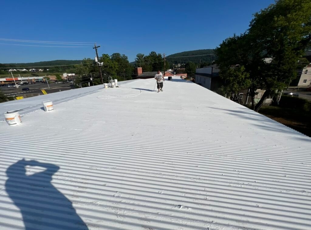 An Anthracite Roofing Systems crew member applying Mule-Hide white acrylic coating to a ribbed metal commercial roof, highlighting leak prevention agreements in Northeastern Pennsylvania (NEPA).